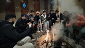 Eine Gruppe von Menschen sitzt an einem Feuer in einem Raum mit Holztüren und einer an der Decke hängenden Gebetsfahne.