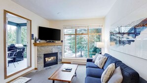 Un salon avec une cheminée, un téléviseur, un canapé et une grande fenêtre donnant sur les arbres.