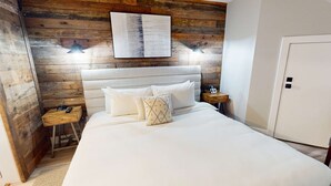 Ein ordentlich bezogenes Bett mit weißen Leinen und Kissen, ein Holz-Kopfbrett und ein Wandbild darüber. Auf dem Nachttisch einer sind eine Lampe und ein Telefon zu sehen.