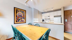 Ein Essbereich mit Holztisch und türkisfarbenen Stühlen, eine Küche mit weißen Schränken und einer Mikrowelle, sowie ein gerahmtes Kunstwerk an der Wand.