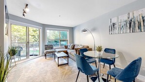Ein modernes Wohnzimmer mit einem Glastisch, blauen Stühlen, einem Sofa und einem großen Fenster mit Blick auf Bäume.