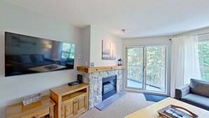 Una sala de estar con chimenea de piedra, un televisor de pantalla plana grande montado en la pared y un ventanal con vistas a árboles.