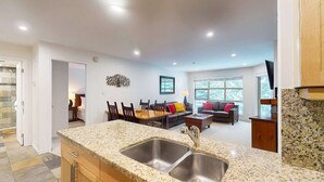 Una cocina moderna con encimeras de granito y lavamanos de acero inoxidable, un comedor con mesa y sillas de madera, una sala de estar con sofá y mesa de centro, y un amplio ventanal con vistas a árboles.