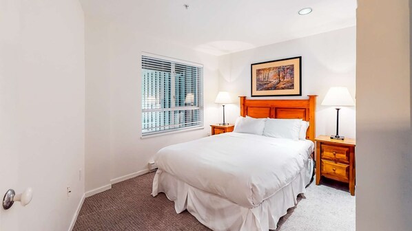 A hotel room with a large bed, two bedside tables with lamps, a window with blinds, and a painting on the wall. - Whistler - Town Plaza 209 (Whistler)