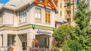 Rakennuksessa on kyltti, jossa lukee ”Whistler Peak Lodge”. Siinä on selkeästi erottuva sisäänkäynti katetulla kuistilla, ulkona on pysäköitynä polkupyörä ja etualalla suuri ikivihreä puu.