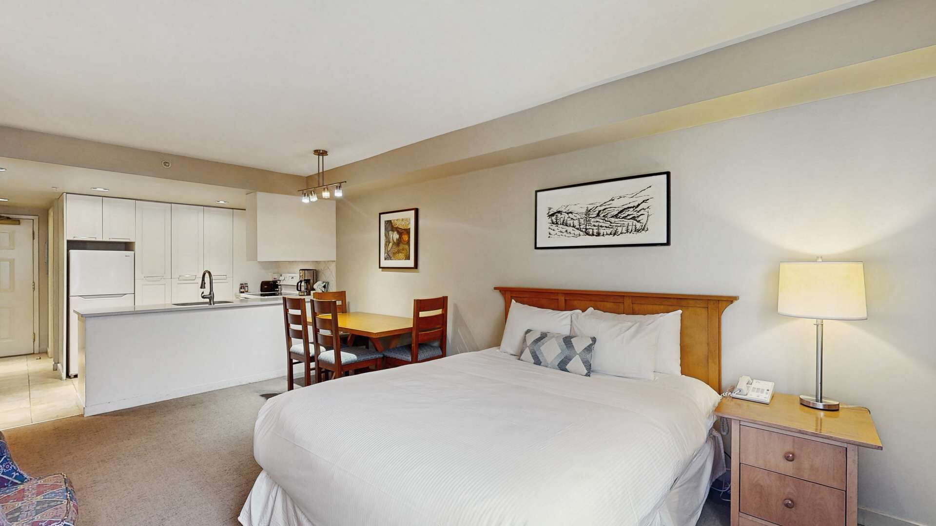 A hotel room with a bed, bedside table, lamp, desk, chair, and kitchenette.