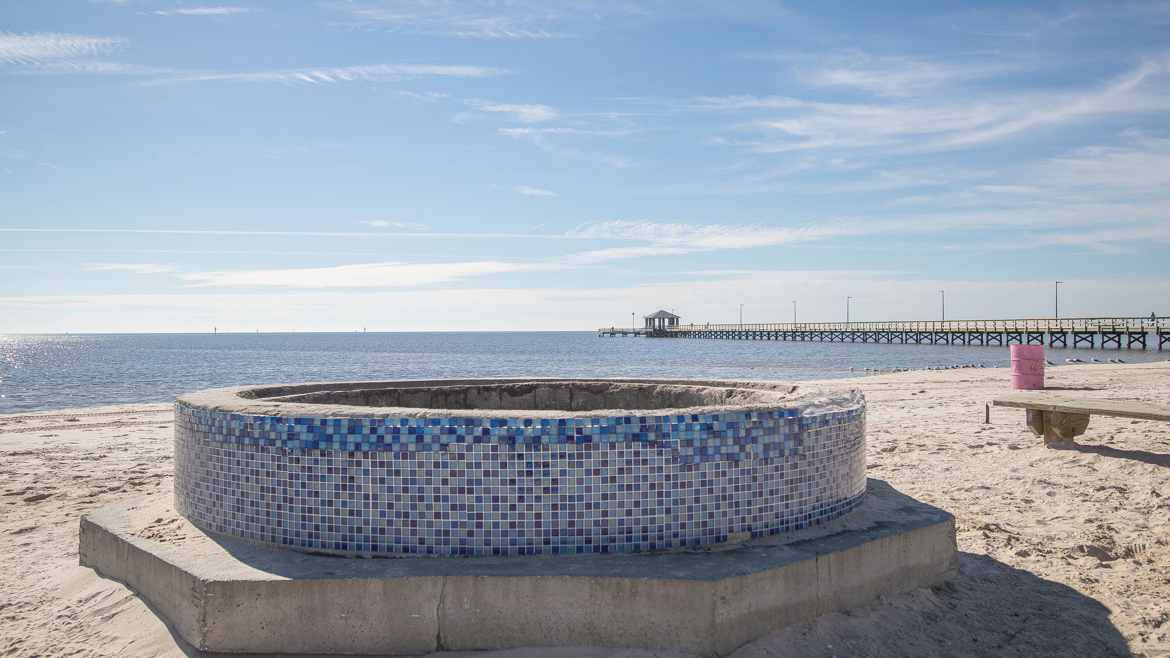 Ein Strand mit einer mosaikverzierten Struktur, einer hölzernen Bank und einem Pier im Hintergrund.