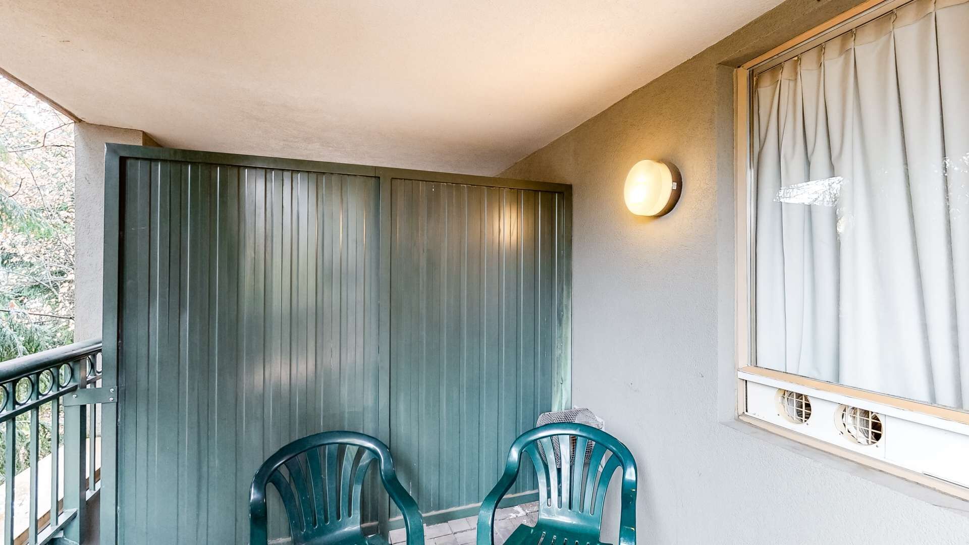 A balcony with two green plastic chairs, a metal railing, a light fixture, and a window with curtains.