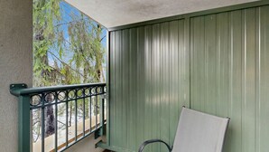 Un balcón con barandilla de metal, una silla y vistas a árboles y al cielo.