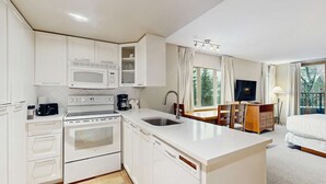 Una cocina moderna con microondas, estufa y lavavajillas, un comedor con mesa y sillas, y un dormitorio con cama y televisor.