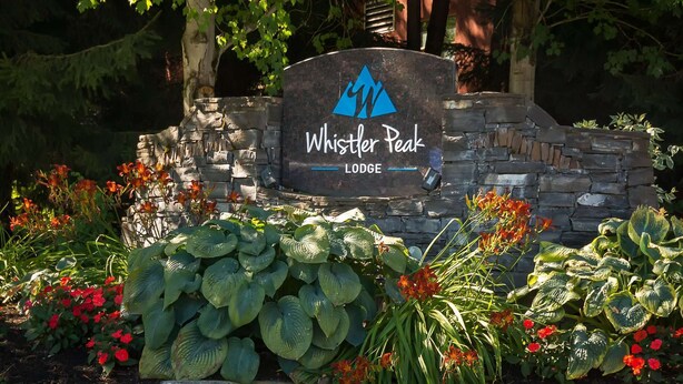 Een bord voor Whistler Peak Lodge, omringd door weelderige begroeiing en bloeiende planten.