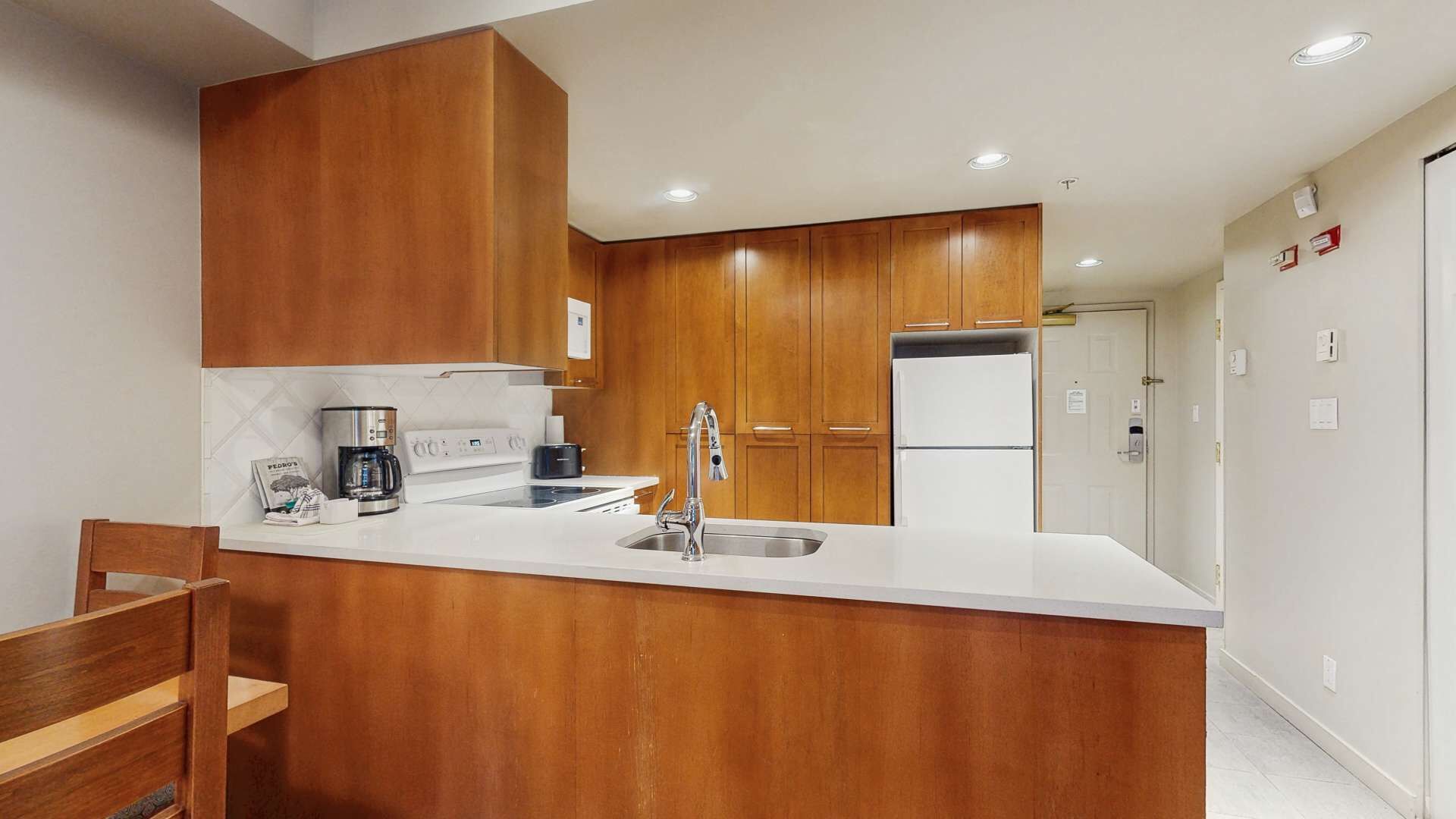 A modern kitchen with wooden cabinets, a white countertop, a stainless steel sink, a coffee maker, and a refrigerator.