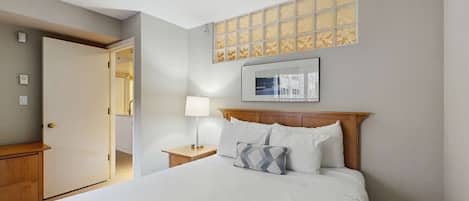 Quarto de hotel com cama, mesa de cabeceira, luminária, porta e janela com vista.