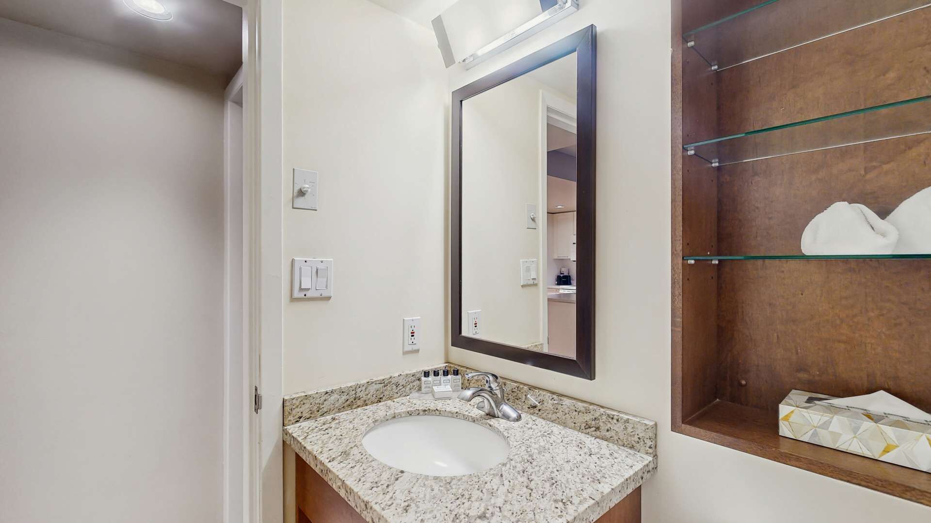 Un baño con lavamanos de encimera de granito, un espejo grande y una estantería de vidrio con artículos de aseo.