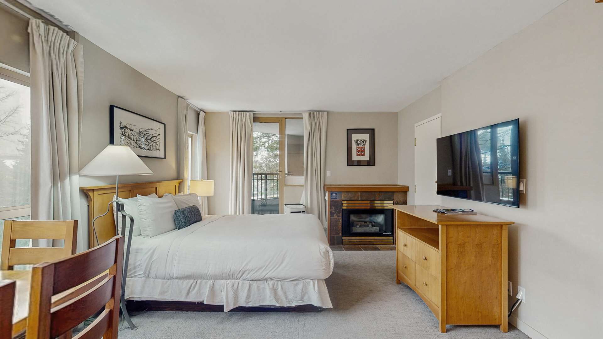 A hotel room with a bed, bedside tables, a TV, a fireplace, and a window with curtains.