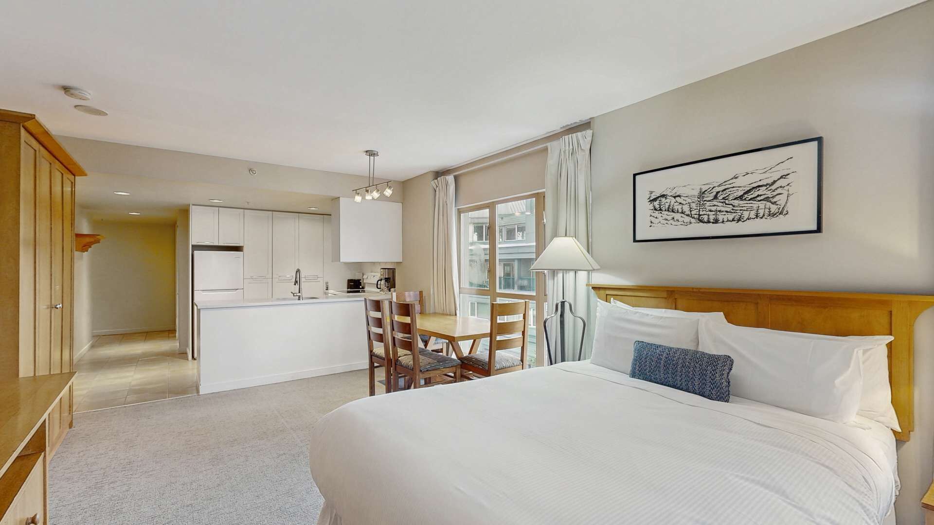 A hotel room with a bed, a desk, a chair, a window, and a kitchen area.