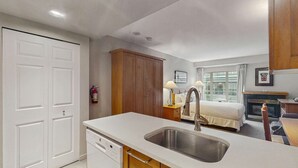Habitación de hotel compacta con kitchenette, cama, mesita de noche con lámparas, chimenea y un amplio ventanal.