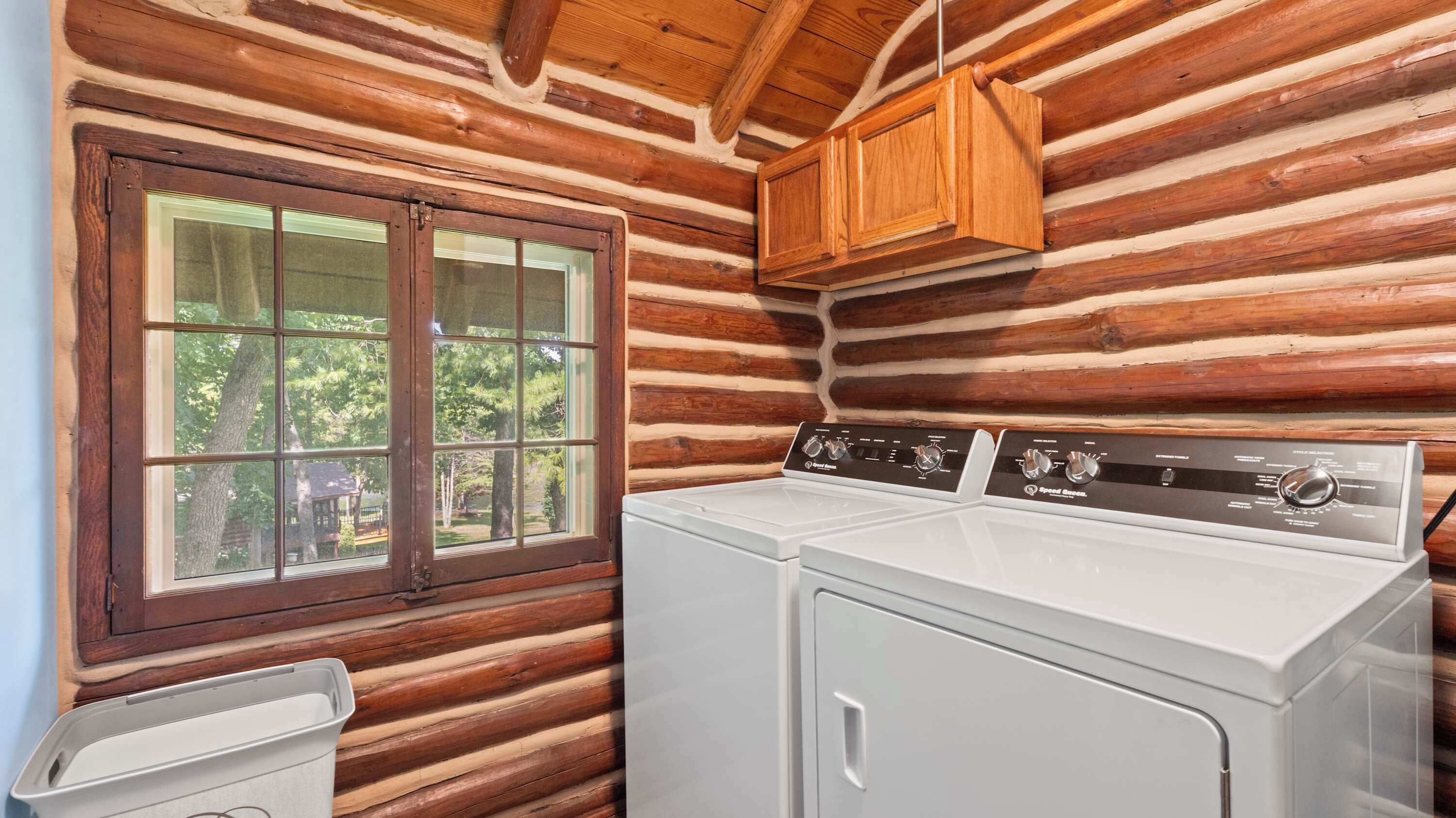 A laundry room with a washer and dryer, wooden walls, a window, and a wooden cabinet.