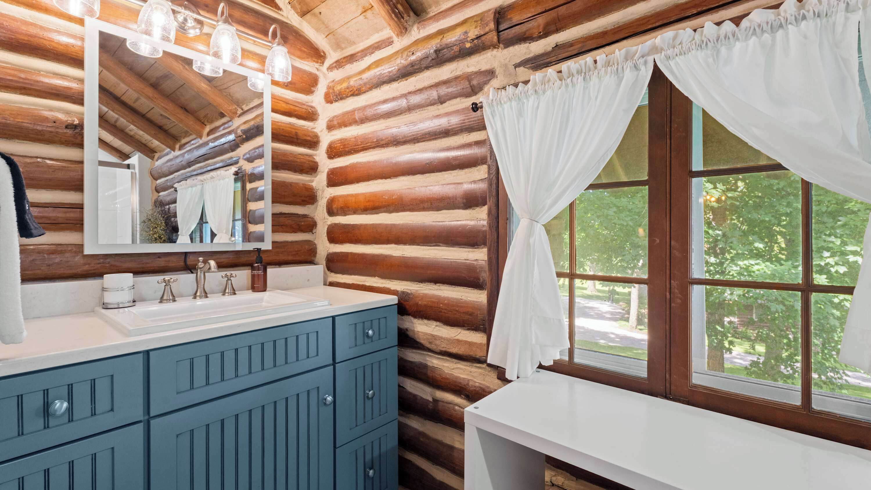 A log cabin bathroom with a white vanity, a mirror, and a window with white curtains.