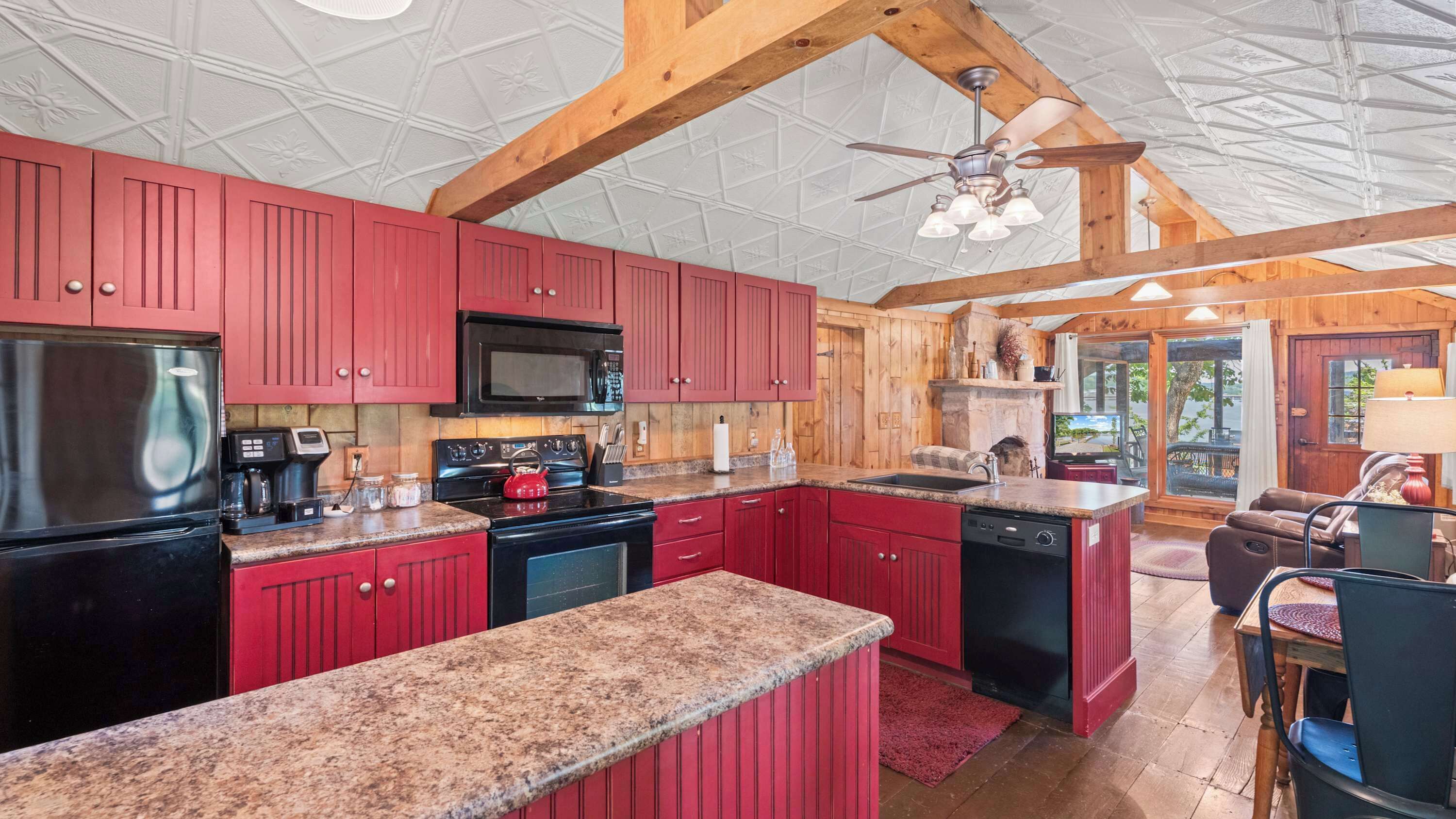 A kitchen with red cabinets, a black refrigerator, a microwave, and a stove. There is a ceiling fan and a stone fireplace.