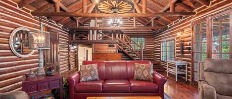 A log cabin interior with a leather sofa, wooden staircase, and a large wooden wheel chandelier.