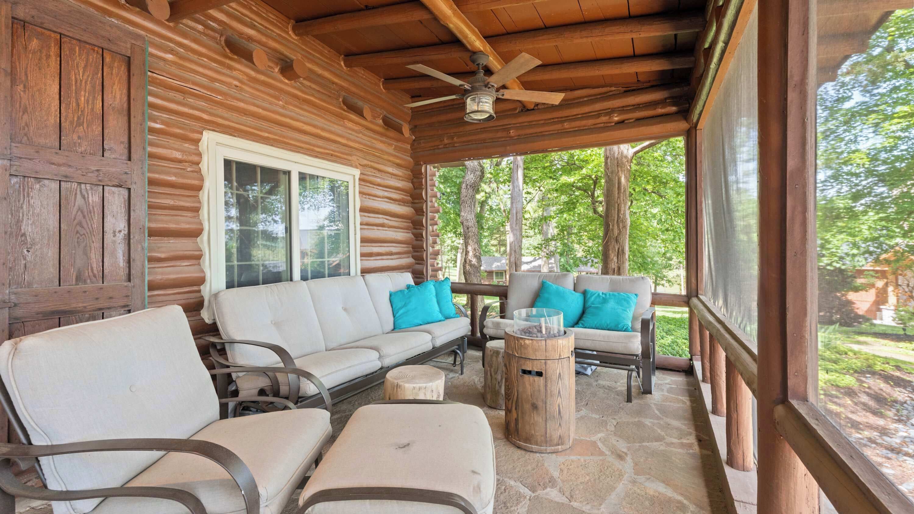 A log cabin with a porch, furnished with a sofa and chairs, a ceiling fan, and a view of the surrounding trees.