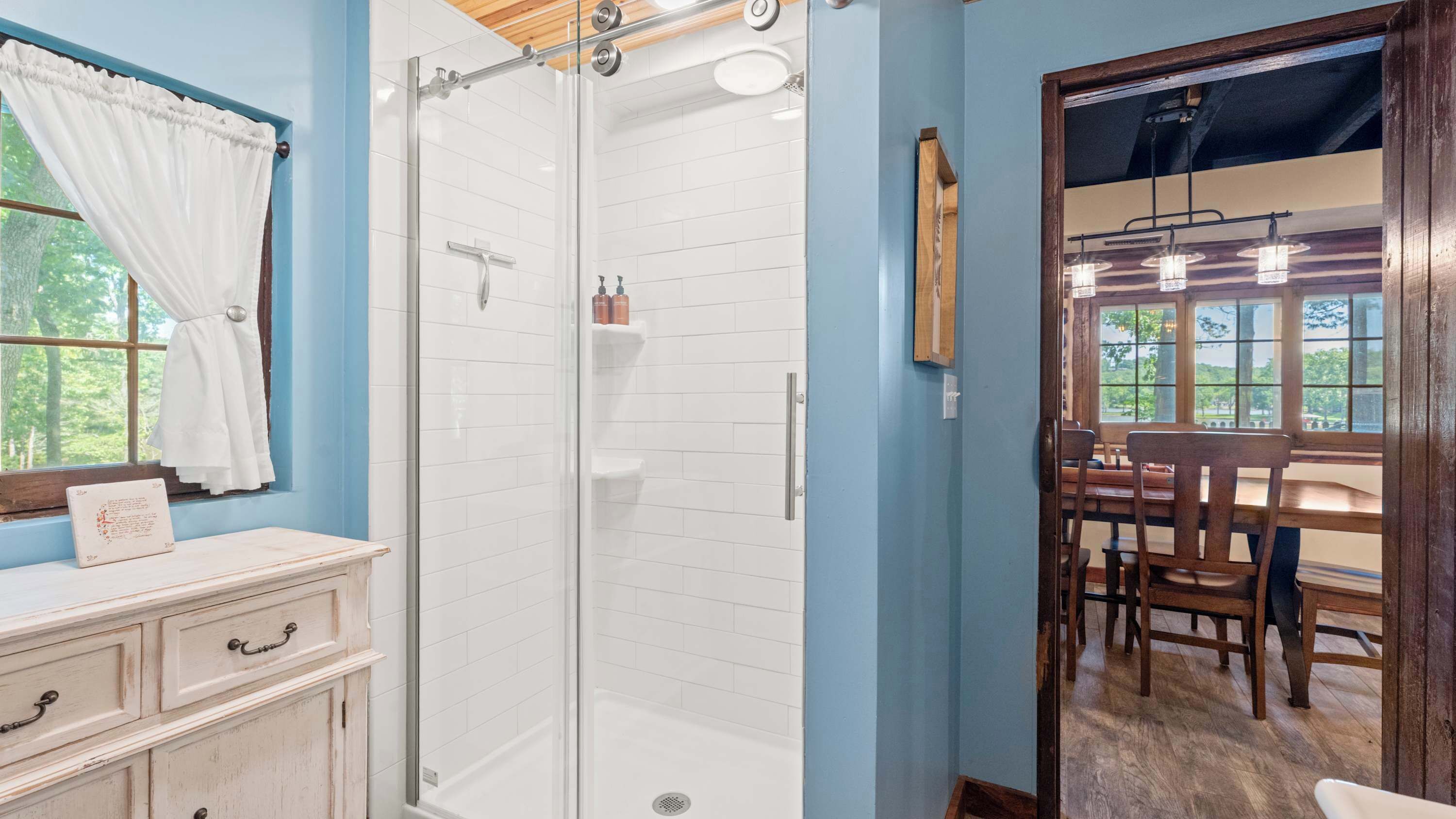 A bathroom with a glass shower enclosure, white tiled walls, a wooden vanity with drawers, and a window with a view of trees.