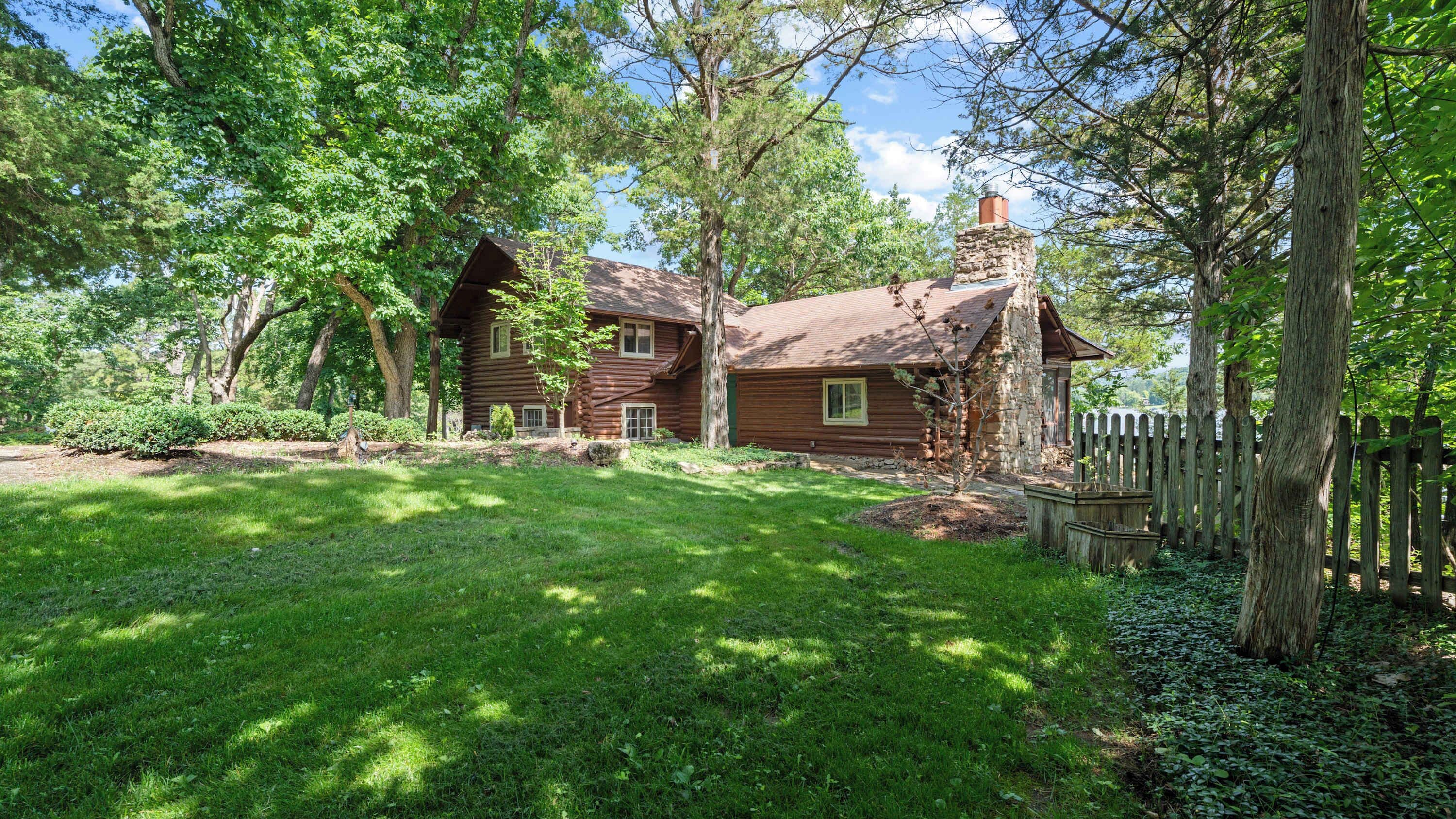 A rustic log cabin with a stone chimney, surrounded by lush greenery and a wooden fence.