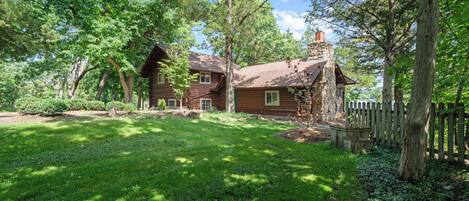 A rustic log cabin with a stone chimney, surrounded by lush greenery and a wooden fence.