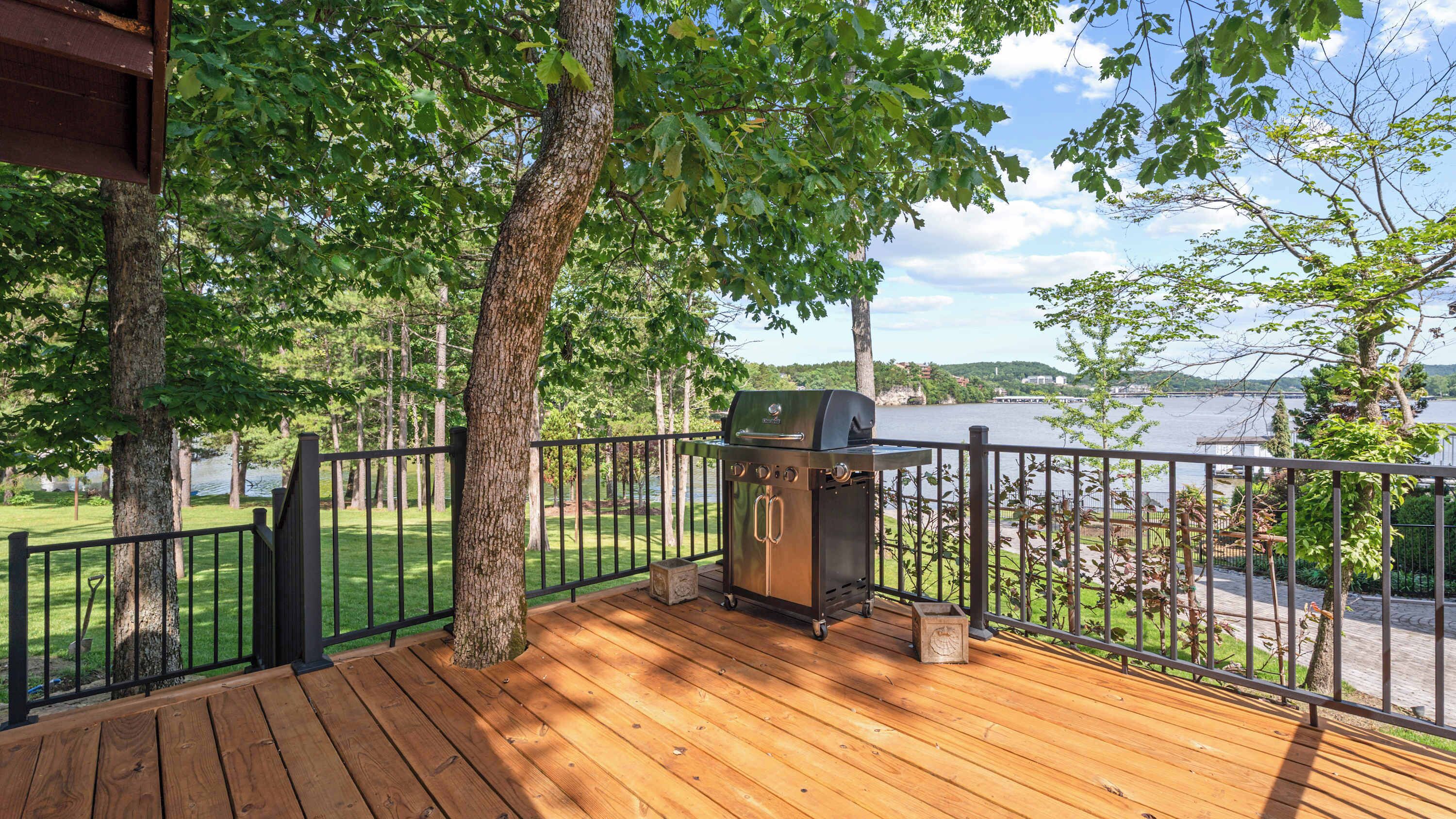 A wooden deck with a barbecue grill, surrounded by a metal railing, overlooking a body of water and greenery.