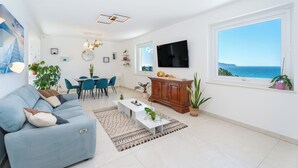Una sala moderna con sofá, mesa de centro y comedor. Hay un televisor montado en la pared y un ventanal con vistas al mar.