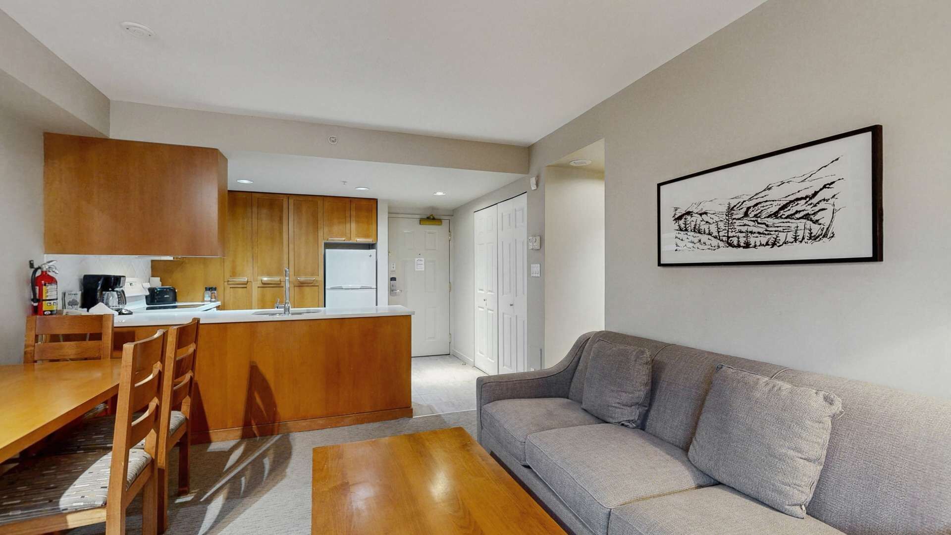 A modern kitchen with wooden cabinets, a dining table with chairs, a grey sofa, and a framed artwork on the wall.