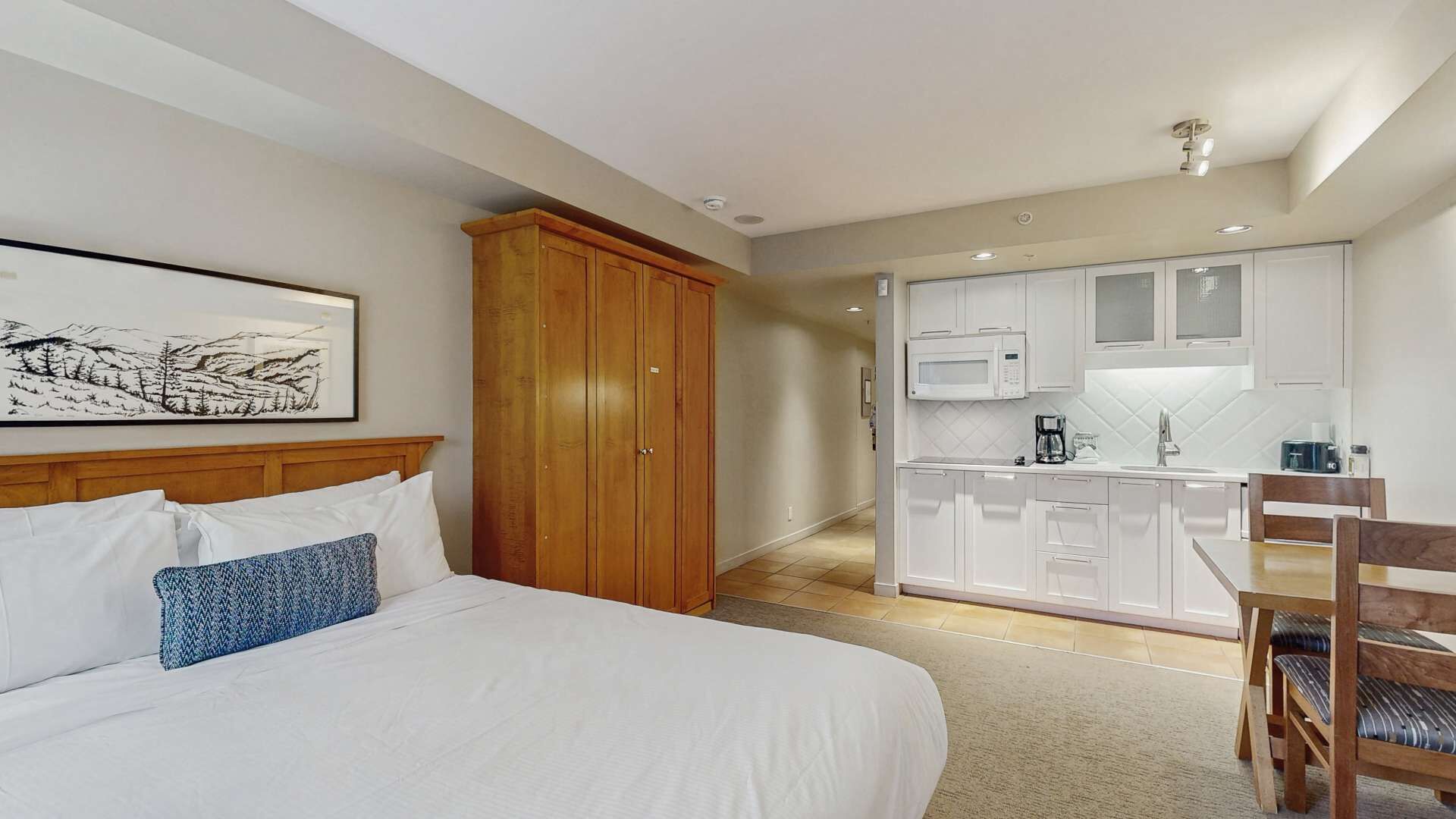 A hotel room with a bed, a wooden wardrobe, a kitchenette, and a dining area.