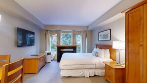 Ett hotellrum med en säng, ett skrivbord, en stol, en TV, en öppen spis och ett fönster med gardiner.