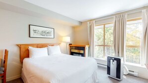 Una habitación de hotel con una cama grande, una mesita de noche con lámpara, un pequeño calentador y una ventana con cortinas.