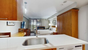 Ein kleines Hotelzimmer mit Küchenzeile, einem Bett, einem Schreibtisch und einem Fenster mit Vorhängen.