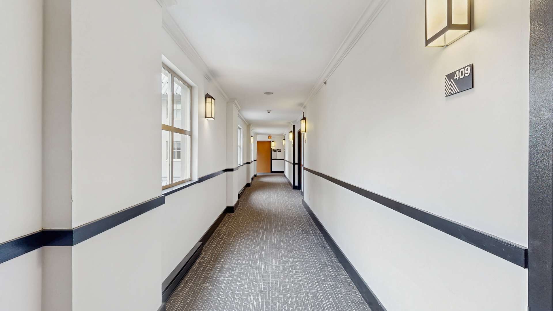Ein Hotelgang mit Teppichboden, weißen Wänden, schwarzer Bordüre und Wandleuchten.