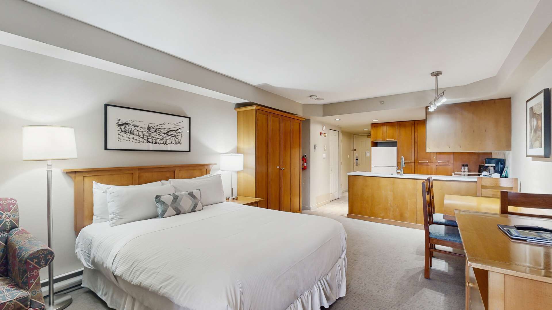 Une chambre d’hôtel comprenant un lit, des lampes de chevet, un bureau, une chaise, une kitchenette et une armoire en bois.