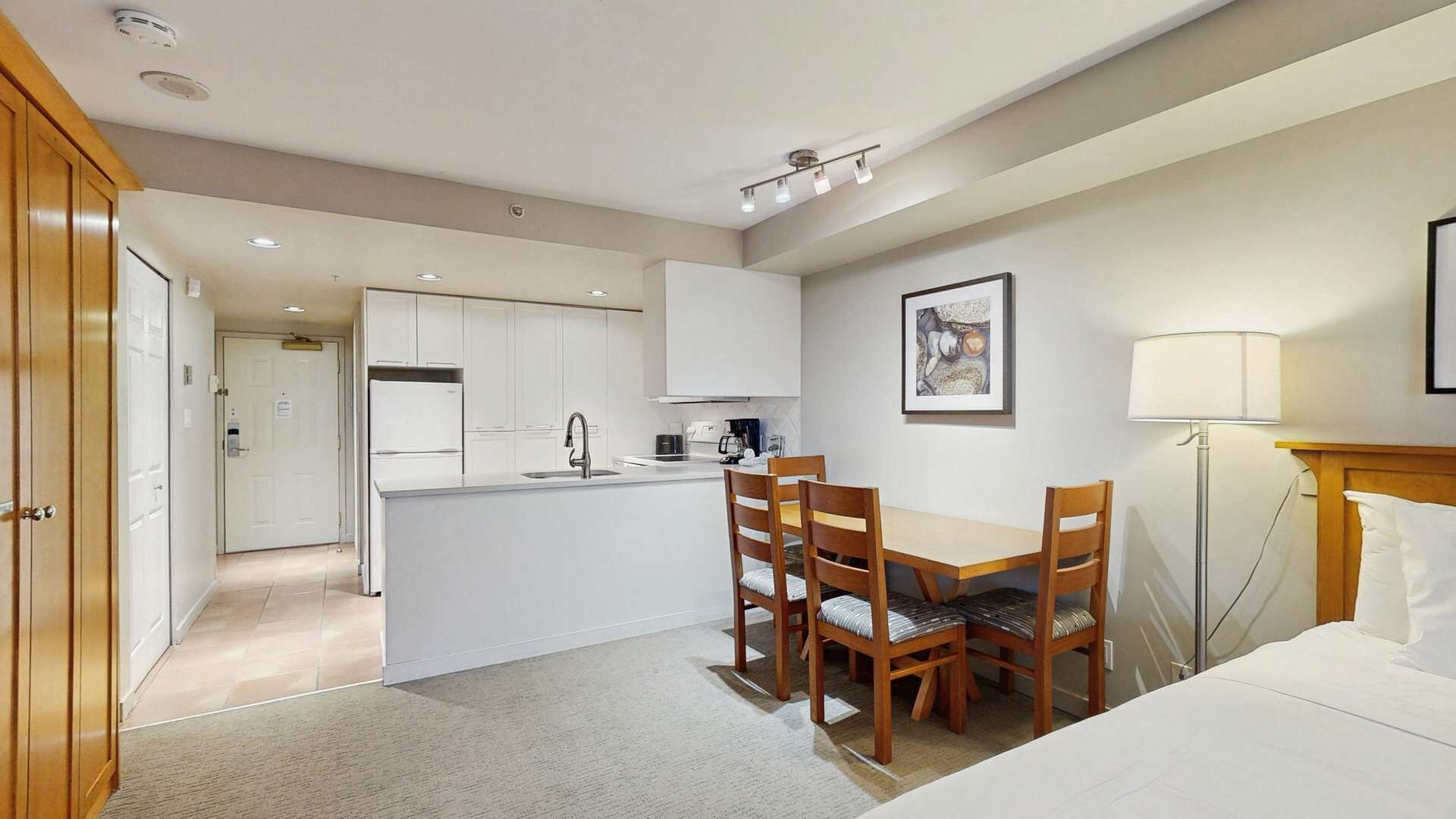 A compact hotel room with a kitchenette, a dining area with two chairs, a bed, and a bedside lamp.
