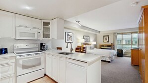 Habitación de hotel compacta con kitchenette, cama, mesita de noche, lámpara y ventana con vistas.