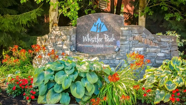 Kivestä valmistettu kyltti, jossa lukee ”Whistler Peak Lodge”, ja sen ympärillä on rehevää kasvillisuutta ja kukkivia kasveja.