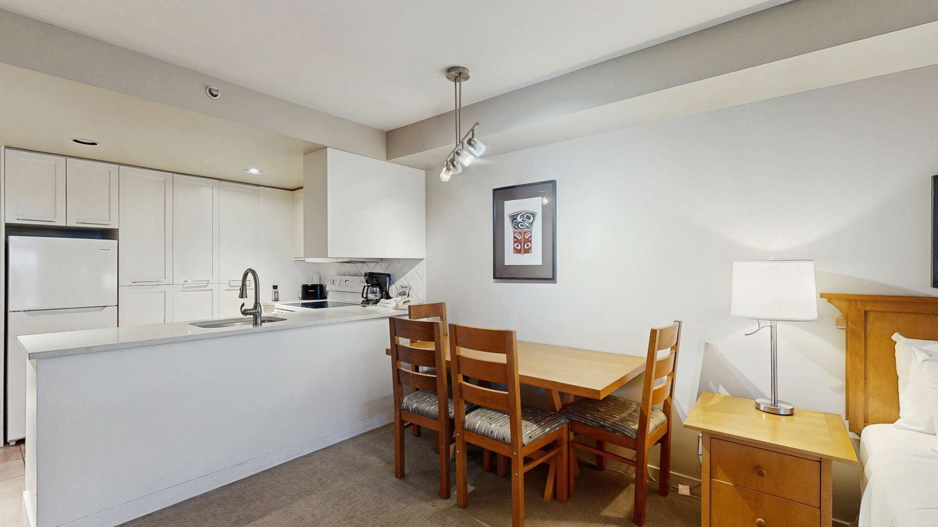 Une cuisine compacte avec des meubles blancs, un coin repas avec des chaises en bois, une petite table et une table de chevet avec une lampe.