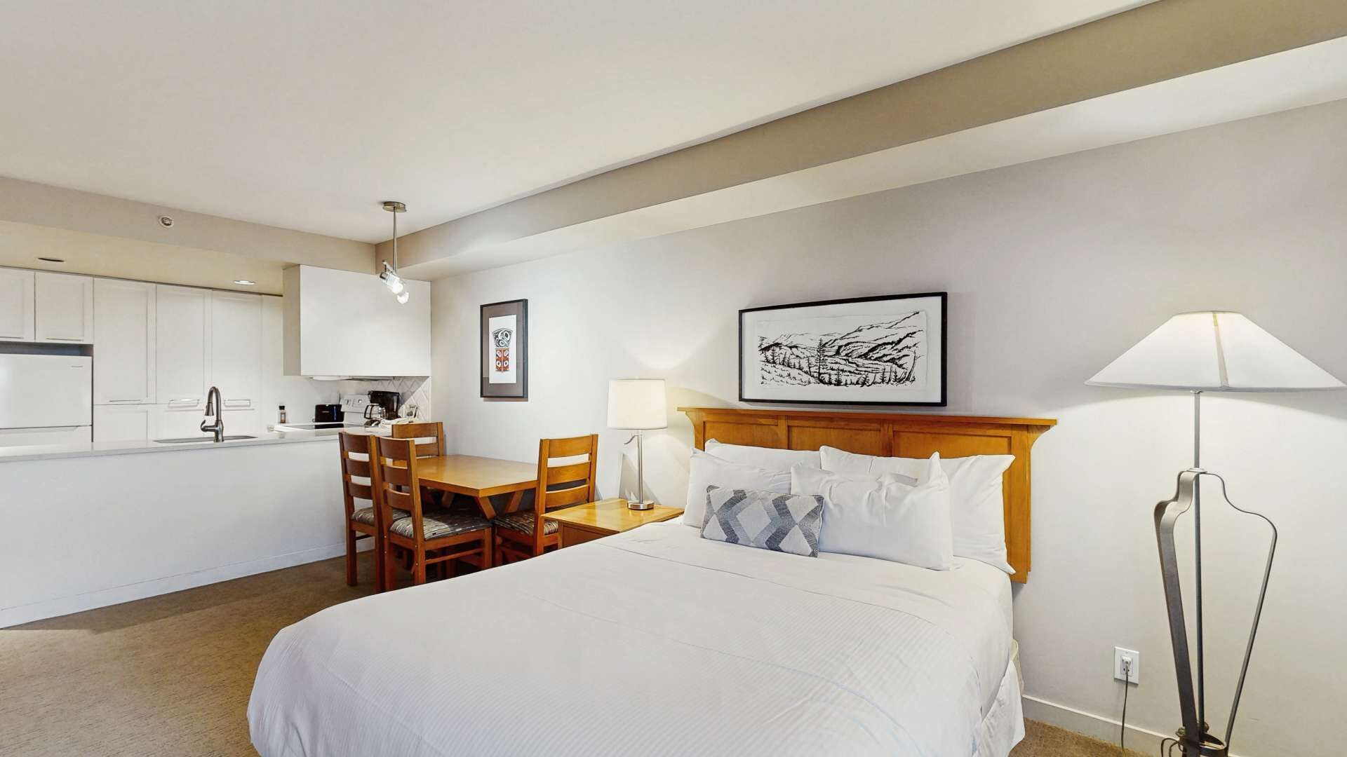 A hotel room with a bed, a desk, a chair, a lamp, and a kitchen area.