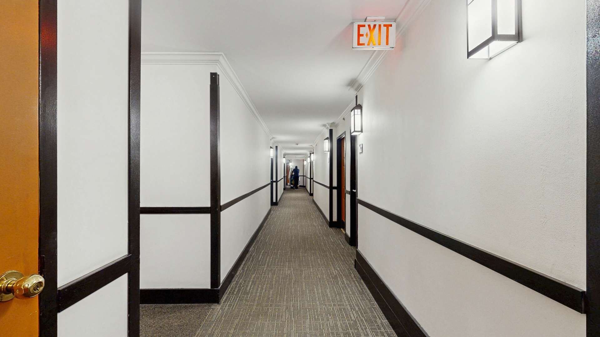 A hotel hallway with a carpeted floor, white walls, black trim, and an exit sign.