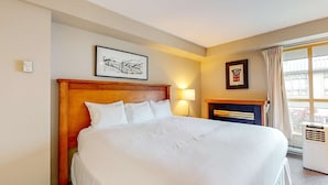 Habitación de hotel con una cama grande, cabecera, chimenea, ventana con cortinas y dos cuadros en la pared.