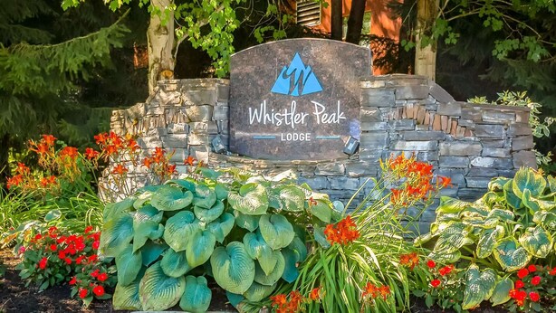Un letrero de piedra con un logotipo y el texto 'Whistler Peak Lodge', rodeado de vegetación exuberante y flores.