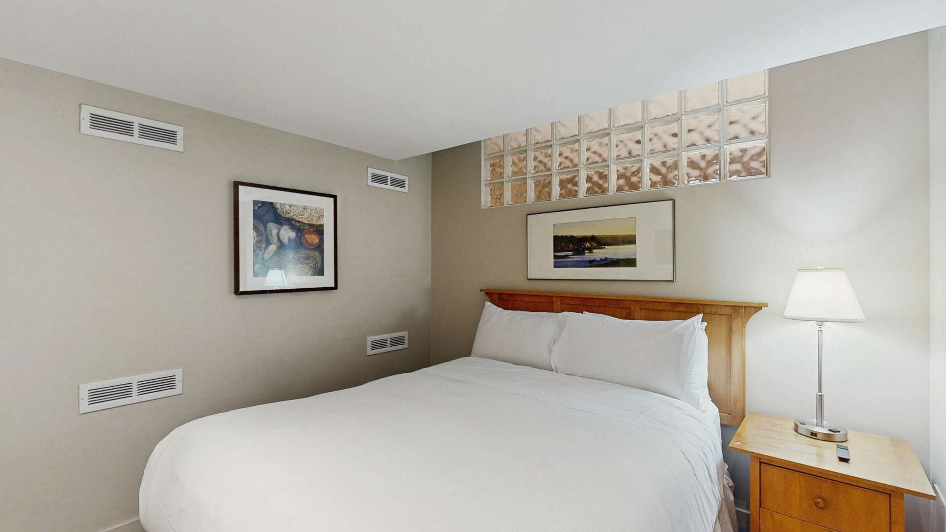 Una habitación de hotel con cama, mesita de noche, lámpara y dos rejillas de ventilación en la pared.