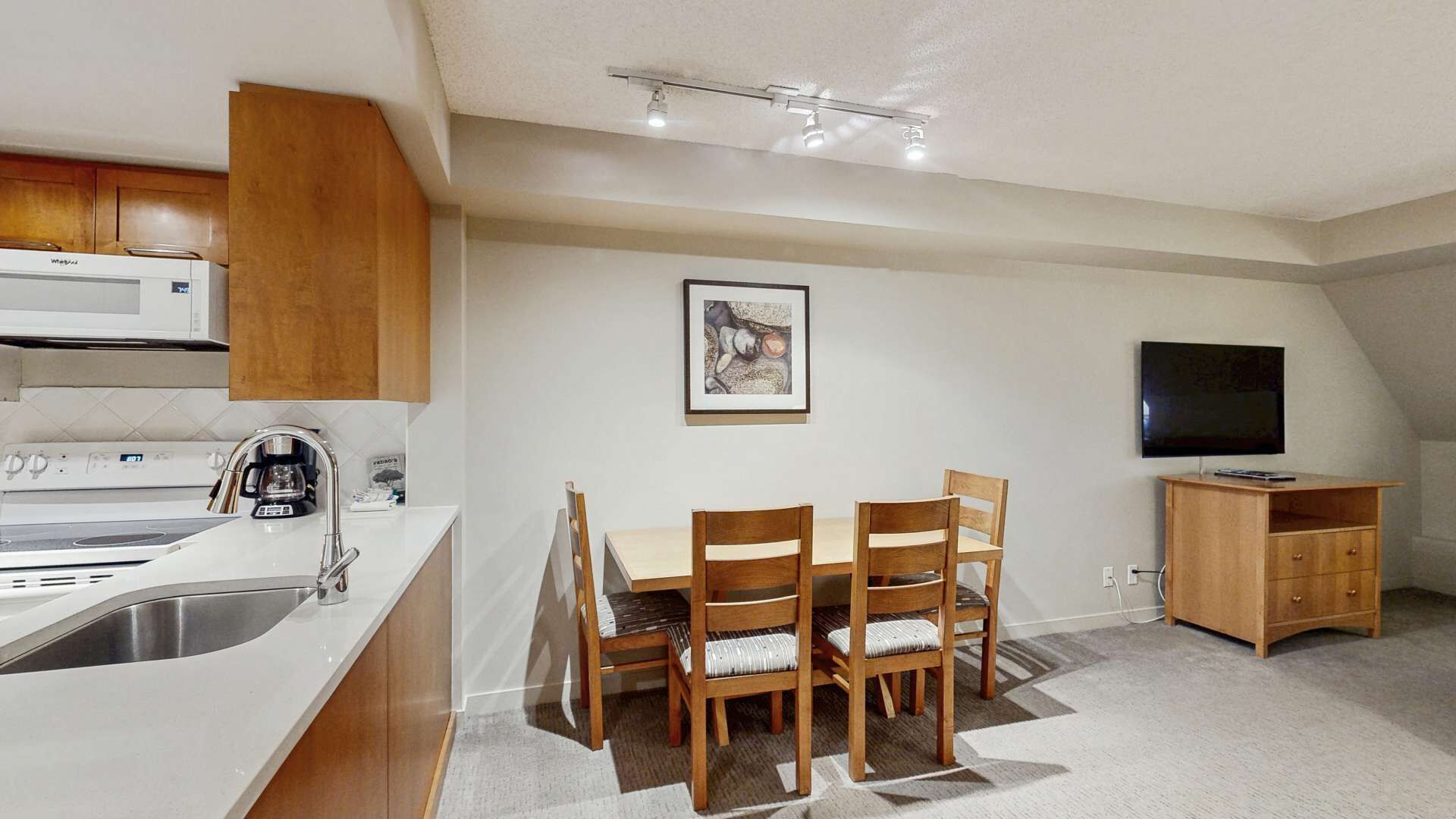 Ett kompakt kök med mikrovågsugn, spis och diskho, en matplats med bord och stolar samt en tv placerad på ett träställ.