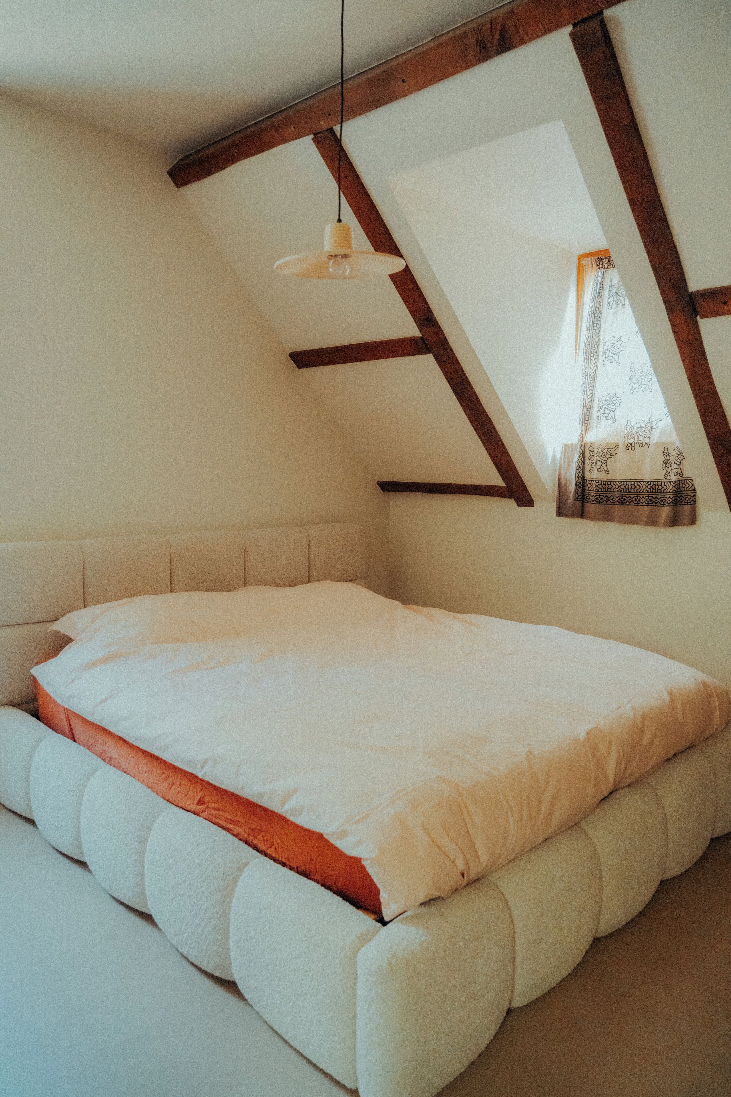 Un dormitorio con techo inclinado, cama con ropa de cama blanca y naranja, y una lámpara colgante.