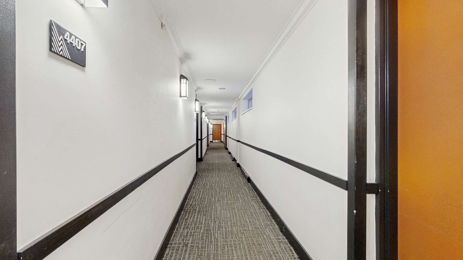 Un pasillo de hotel con piso alfombrado, paredes blancas y una puerta a la derecha.